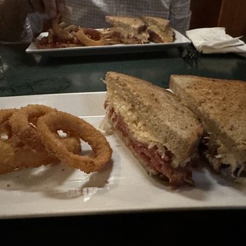 Packards - Onion rings and corned beef Reuben. - A rectangular white plate contains a serving of onion rings and a half-eaten Reuben sandwich with toasted bread, corned beef, melted cheese, and possibly sauerkraut. The plate is on a dark table in a dimly lit setting, with a glass, napkins, and a ketchup bottle visible nearby. In the background, another plate with food and a person’s arm are partially visible. - Pubs Near Me - Northampton, Massachusetts