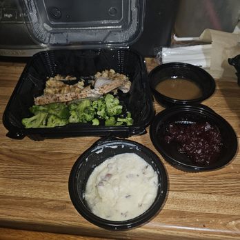 Denny's - This is what I received x 4 still not the same amount that was in the ad - The image shows a takeout meal with four black plastic containers placed on a wooden counter. One container holds steamed broccoli and a piece of cooked meat. The other three containers contain mashed potatoes with gravy, cranberry sauce, and additional gravy or sauce. The background includes a napkin holder, a metal container, and part of a kitchen appliance, indicating a casual dining or takeout setting. - Diners Near Me - Bastrop, Texas
