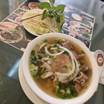 Pho Bang Restaurant - A bowl of Vietnamese pho with beef slices, chopped green onions, and herbs in a clear broth. Accompanying garnishes include bean sprouts, lime wedges, and fresh basil leaves on a plate. The table has a menu with images of dishes, indicating a Vietnamese restaurant setting. - Vietnamese Near Me - Flushing, New York