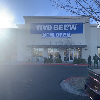 Five Below - The image shows the front exterior of a Five Below retail store with a prominent sign displaying the store's name and an additional banner indicating it is now open. The storefront has large glass windows and doors, with a few people walking toward the entrance. The scene is set in a parking lot with some cars visible, and a tree with bare branches is seen on the right side of the image. The sky is clear and sunny. - Discount Store Near Me - Albuquerque, New Mexico