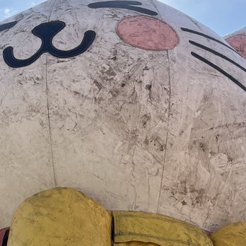 Funbox - A weathered sculpture of a cartoon cat's face with a yellow body, pink nose, and black whiskers, set outdoors against a blue sky. - Kids Activities Near Me - Lombard, Illinois