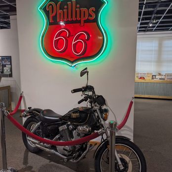 Route 66 State Park - A black vintage motorcycle with chrome details is displayed indoors, surrounded by a red velvet rope barrier. Behind the motorcycle, a neon sign features the Route 66 shield with the Phillips logo and the number 66 illuminated in red and green. The setting appears to be an exhibit or museum display at Route 66 State Park, with a neutral wall and additional interior elements visible in the background. - Parks Near Me - Eureka, Missouri
