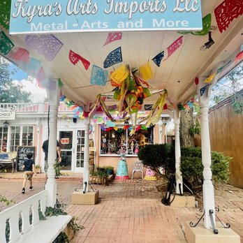 La Choco - The image shows the storefront sign for Kyra's Arts Imports LLC, which offers metal arts and more. The sign is hanging above a decorated outdoor patio area with colorful papel picado banners and festive decorations hanging from the ceiling. The patio has white columns, a brick pathway, and some greenery, with a view of neighboring shops and people walking nearby. - Desserts Near Me - Albuquerque, New Mexico