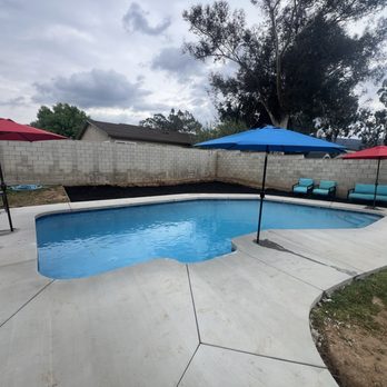Cerda Pool Construction - The image shows a backyard swimming pool with a concrete deck surrounding it. There are three umbrellas—two red and one blue—providing shade over the seating area, which includes three chairs and a bench. The pool is filled with water, and the area is enclosed by a brick wall. The background includes trees, a cloudy sky, and neighboring houses, indicating a residential setting. - Pool & Hot Tub Service Near Me