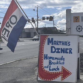 Martha's Diner - The sign by the highway. - A sidewalk sign for Martha's Diner advertises homemade breakfast and lunch, with a red arrow pointing right. An American flag is attached nearby, and the background shows a street with cars and utility poles. - American Near Me - Ellsworth, Maine