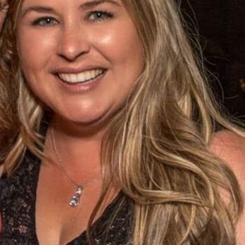 Glamor Cuts - A woman with long, wavy blonde hair and a black lace top is smiling at a social gathering, wearing a necklace with a pendant. - Hair Salons Near Me - Concord, California
