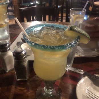 The Common Man Windham - A margarita cocktail with ice, served in a glass with a salted rim and a lime wedge garnish. The drink is yellowish in color and is placed on a wooden table in a restaurant or bar setting. There are other drinks and condiments visible in the background. - New American Near Me - Windham, New Hampshire