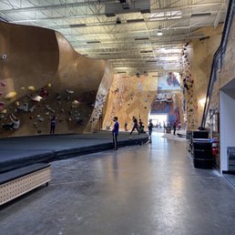 A spacious indoor rock climbing gym features multiple climbing walls with different angles and holds, with climbers and belayers present.