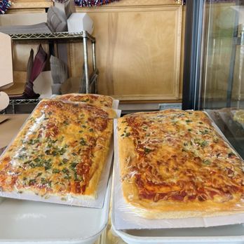 Mazzetti's Bakery - Foccacia - Uncooked rectangular pizzas with cheese and toppings are placed on trays inside a bakery display case, with a decorated bakery interior in the background. - Bakeries Near Me - Pacifica, California
