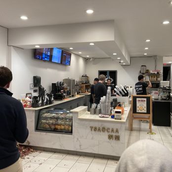 Tsaocaa - Busy behind the counter - The image shows the interior of Tsaocaa, a bubble tea shop in Pittsburgh, PA. The space includes a counter with staff preparing drinks, self-order kiosks, and a display case with baked goods. Customers are standing in line or waiting, and the shop has a clean, modern design with white walls and ceiling lights. The storefront is not visible. - Bubble Tea Near Me - Pittsburgh, Pennsylvania