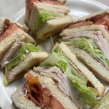 Village Grill - Club - A plate contains multiple small sandwiches made with white bread, filled with lettuce, sliced turkey, tomato, and bacon. The sandwiches are cut into halves or quarters, arranged on a white plate, with a pickle spear visible at the edge of the plate. The image focuses on the sandwiches, showing fresh ingredients and a simple presentation. - Diners Near Me - Tehachapi, California