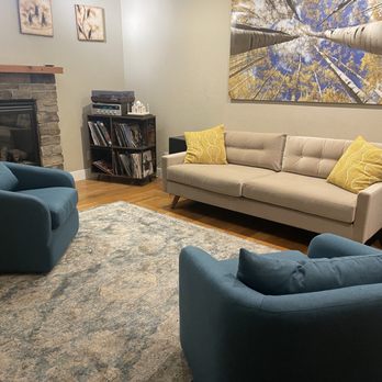 Joybird - The image shows a living room with a stone fireplace on the left, topped with a wooden mantel. Two blue armchairs are positioned facing each other, with a beige sofa against the right wall, decorated with two yellow patterned pillows. Above the sofa, there is a large wall art piece depicting a view of tall trees from below. A black shelving unit with vinyl records and a stereo system is placed near the fireplace. The room has a wooden floor and a patterned area rug covering part of the floor. - Rugs Near Me - Denver, Colorado
