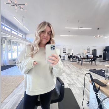 Arrow & Mane - A woman with wavy blonde hair in a white sweatshirt and black leggings is taking a mirror selfie in a hair salon with contemporary lighting and decor. - Hair Extensions Near Me - Tracy, California