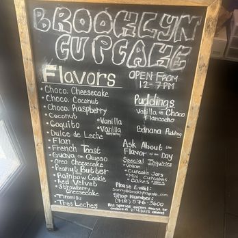 Brooklyn Cupcake - A chalkboard sign with a wooden frame is positioned indoors, listing cupcake flavors such as cheesecake, coconut, raspberry, and more. It also mentions pudding flavors like vanilla and choco, and provides business hours from 12 PM to 7 PM. The sign includes contact information and notes about special orders and inquiries, indicating it is for Brooklyn Cupcake, a bakery offering diverse cupcake and pudding flavors. - Cupcakes Near Me - Brooklyn, New York
