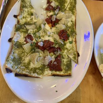 M'tucci's Italian - Sun dried tomatoes and pesto flatbread pizza - A rectangular flatbread pizza features a thin crust topped with green pesto sauce, sun-dried tomatoes, artichoke hearts, and chopped herbs. The pizza is served on a white oval plate, with some toppings scattered around the edges. The dish appears to be part of a meal at an Italian restaurant. - Italian Near Me - Albuquerque, New Mexico