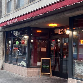 Red's Place - The photo depicts the storefront of Red's Place, a bar and pub located in Ithaca, NY. The establishment has a red awning with the name 'red's place' visible on it, and large glass windows revealing the interior lighting and decor. A chalkboard sign is placed outside the entrance, and the building has a brick facade with multiple windows above. The sidewalk in front is empty, and the overall scene suggests an urban setting. - New American Near Me - Ithaca, New York