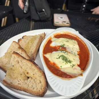 The Corner Store - Shakshuka - Shakshuka with melted cheese and herbs is served on a white dish, accompanied by three slices of toasted bread, on a black outdoor table. - Coffee & Tea Near Me - Crown Heights, New York