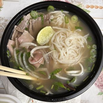 Pho King Orem - Number 8 pho - A bowl of Vietnamese pho contains rice noodles, thinly sliced beef, chopped green onions, and a lime wedge. The broth is clear and light, with visible green herbs and vegetables. The bowl is black, placed on a floral-patterned tablecloth, with chopsticks resting on the side. - Vietnamese Near Me - Orem, Utah
