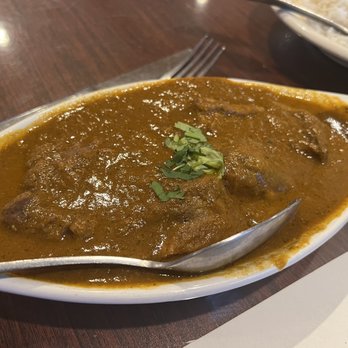 Namaste - Lamb Curry - A white, oval-shaped ceramic dish holds lamb curry with a rich, brown sauce and tender pieces of lamb. The dish is garnished with a small amount of chopped herbs on top. The curry is served on a wooden table, with a metal spoon resting inside the dish. In the background, part of a plate of rice and a fork are visible. - Indian Near Me - Rio Rancho, New Mexico