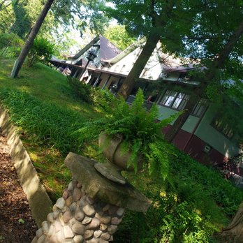 Lakeside Inn - A garden with a stone pedestal and planter filled with ferns, set among trees and plants, with a house in the background. - Bed & Breakfast Near Me - Lakeside, Michigan