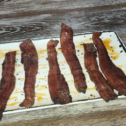 Six strips of cooked bacon are arranged in a row on a rectangular white plate, which has sauce drizzles underneath and around the bacon. The plate is on a wooden table with a textured surface. In the background, part of a person's arm and torso are visible, with the person wearing a light pink sweater and a bracelet. The setting appears to be a casual dining environment.