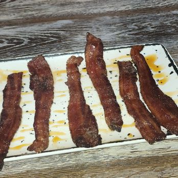 Maple + Bacon - Six strips of cooked bacon are arranged in a row on a rectangular white plate, which has sauce drizzles underneath and around the bacon. The plate is on a wooden table with a textured surface. In the background, part of a person's arm and torso are visible, with the person wearing a light pink sweater and a bracelet. The setting appears to be a casual dining environment. - Breakfast & Brunch Near Me - Valparaiso, Indiana