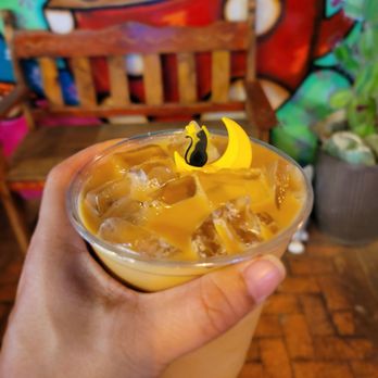 Blackbird Coffee House - Jimmy Crack Corn.. the cute cat on a crescent moon stirring stick. - A cold drink with ice, topped with a Halloween-themed decoration of a black cat on a yellow crescent moon, held in front of a colorful mural. - Coffee & Tea Near Me - Albuquerque, New Mexico