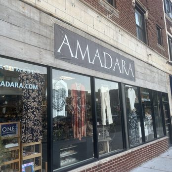 AMADARA - The storefront ! - The image shows the exterior of AMADARA, a gift shop located in Chicago, IL. The storefront has large glass windows that reveal mannequins dressed in handmade clothing and jewelry inside. A black and white sign with the shop's name, 'AMADARA', is mounted above the windows. The building's facade includes a brick section above and a concrete or stone section below the sign. The sidewalk in front is visible, and the shop appears to be open during daytime. - Arts & Crafts Near Me - Chicago, Illinois