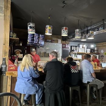 Bald Buddha Brewing - Bar scene - The image shows the interior of Bald Buddha Brewing in Springfield, IL, with customers seated on bar stools around a wooden bar counter. Some patrons are using laptops, while others are engaged in conversation. The bar area features a digital menu screen, brewing supplies, and various decorations, including hanging lights with patterned shades. The setting appears casual and lively, with a focus on craft beer and brewing culture. - Bars Near Me - Springfield, Illinois