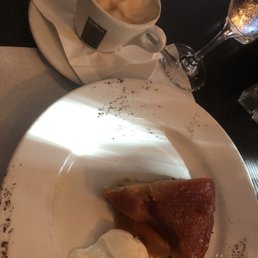 Apple tartin - A dessert apple tartin served on a white plate with a dollop of whipped cream. The plate has a decorative dusting of cocoa powder or cinnamon around the edges. A coffee drink with foam is placed on a saucer with a napkin, and a glass of sparkling wine is also visible on the dark table surface.