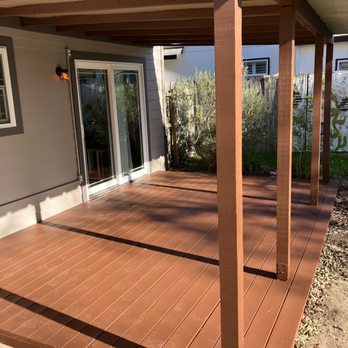 Deck N Cable Railing - A wooden deck with a pergola-style roof is attached to a house with sliding glass doors. The deck is made of brown wood planks and is bordered by a garden with bushes and a wooden fence. The house exterior is gray with white trim, and a small outdoor light is mounted near the sliding doors. The area is illuminated by sunlight, casting shadows on the deck. - Decks & Railing Near Me