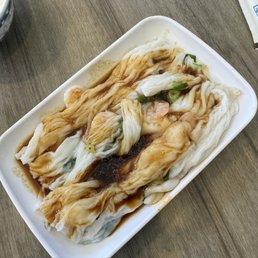 Shrimp Steamed Rice Roll - The image shows a white rectangular plate containing steamed rice rolls filled with shrimp. The rice rolls are drizzled with a dark sauce and garnished with chopped green onions. The plate is placed on a wooden table, with part of a paper napkin visible in the background. The dish is a traditional Cantonese steamed rice roll, often served with soy-based sauce and garnishes.