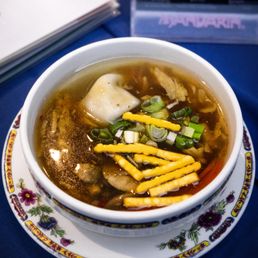 Wonton Soup - A bowl of hot and sour soup containing green onions, shredded cheese, and a wonton, served in a white bowl with a floral-patterned plate underneath. The soup appears clear with visible ingredients floating on the surface. The background includes a menu and a blue tablecloth, indicating a restaurant setting.