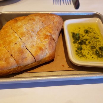 Bonefish Grill - A baked pastry with a golden, flaky crust, sliced into sections, is presented on a metal tray with a piece of parchment paper underneath. Next to it, there is a small rectangular dish containing a green herb-infused oil or sauce with visible herbs and oil separation. The presentation suggests a dish served in a restaurant or cafe setting. - Seafood Near Me - Brookfield, Wisconsin