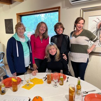 Allan Houser - Five women are gathered around a round dining table in a room with wooden ceiling beams and a large window showing trees outside. The table has orange plates, glasses with drinks, and small bottles. There are pumpkins and dried chili peppers on the table. The women are smiling and dressed casually, with one seated and four standing behind her. The room has a framed artwork on the wall and an air conditioning unit. - Art Galleries Near Me - Santa Fe, New Mexico
