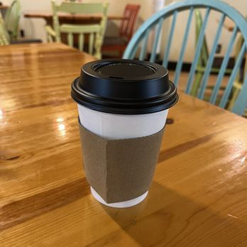 Kickback Cafe - A disposable coffee cup with a black plastic lid and a cardboard sleeve is positioned on a polished wooden table. The background features colorful chairs and a wall with framed pictures, indicating a cafe environment. - Cafes Near Me - Oregon, Wisconsin