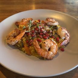 Shrimp risotto - A seafood risotto featuring grilled shrimp arranged on top of a bed of rice mixed with vegetables and herbs. The dish is garnished with microgreens and pomegranate seeds, served on a white, round plate placed on a wooden table. The presentation emphasizes fresh ingredients and colorful toppings.