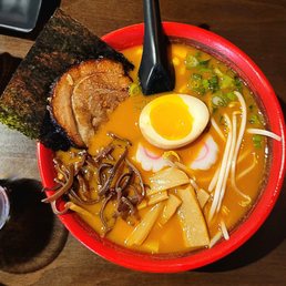 Lava - A bowl of ramen featuring pork belly, fish cake, a lava egg, green onions, bamboo shoots, and seaweed, served in a red bowl.