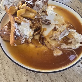 High/Low - French toast - A plate features French toast topped with cooked apple slices, chopped nuts, and a dusting of powdered sugar. The dish is served with syrup, which is visible around the toast and on the plate. The plate is placed on a textured, light-colored table surface, and part of a mug is visible in the background. - Breakfast & Brunch Near Me - Oceanside, California