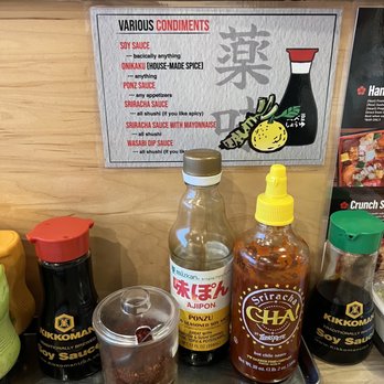Chicago Sushi - September 2022 - The image shows a condiment station with bottles of soy sauce, ponzu, sriracha, and wasabi dip sauces arranged on a tray. A sign above lists various condiments, including soy sauce, onikiku (house-made spice), ponzu sauce, sriracha sauce, sriracha sauce with mayonnaise, and wasabi dip sauce. The sign provides descriptions for each condiment, indicating their typical uses and flavor profiles. - Japanese Near Me - Des Plaines, Illinois