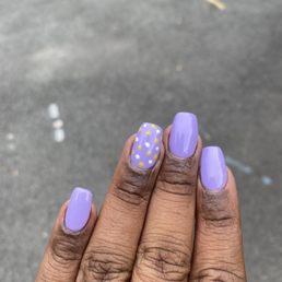 Gel fill - The image shows a hand with four fingers visible, each with lavender-colored gel nails. The middle finger has an accent nail decorated with white and yellow polka dots on a lavender background. The background is an outdoor paved surface, slightly blurred. The nails are well-manicured and glossy.