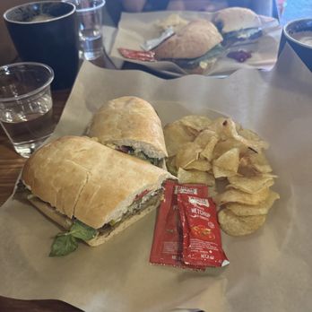 Beyond Cafe - Persian Kotlet Sandwich - A sandwich with bread, vegetables, and possibly meat, served with potato chips and ketchup packets on a paper-lined tray. - Coffee & Tea Near Me - Orleans, Ontario
