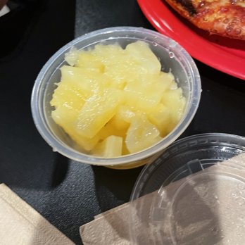 Pizza 9 - Forgotten pineapple - A small, round, clear plastic container is filled with yellow pineapple chunks, positioned on a black table. Part of a red plate with a pizza slice is visible in the background. The container has a transparent lid, which is partially open, and a napkin is placed nearby. The setting appears to be a casual dining environment, possibly a pizzeria. - Pizza Near Me - Grants, New Mexico