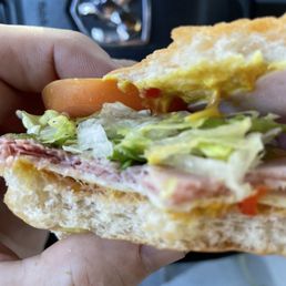 One slice of ham. Or is it 2 paper thin slices - A sandwich with lettuce, tomato, ham, and egg on a bun is held in a person's hand, with a vehicle dashboard visible in the background.