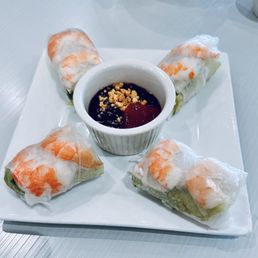 Summer Rolls - Four Vietnamese spring rolls wrapped in translucent rice paper, containing vegetables and shrimp, are arranged around a small white ramekin filled with dipping sauce topped with crushed peanuts. The dish is presented on a white square plate, placed on a light-colored surface.