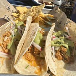 Chicken tacos - Three soft tacos filled with shredded meat, garnished with sliced radishes, shredded lettuce, and drizzled with green sauce. The tacos are served on a tray with tortilla chips and a side of beans in a small metal cup. The setting appears to be a casual dining environment, possibly outdoors or in a restaurant with a relaxed atmosphere.