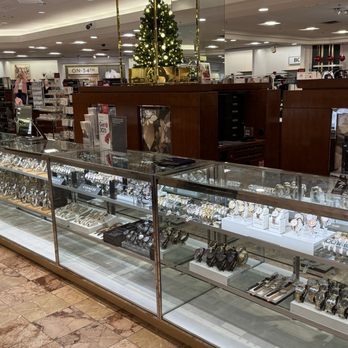 Macy's - A glass jewelry display case showcasing various watches, located in a department store with Christmas decorations and shoppers in the background. - Department Stores Near Me - Albuquerque, New Mexico