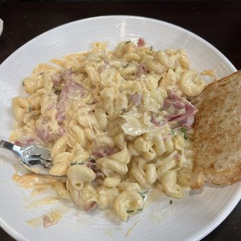 The MacStack Tavern - Reuben Mac n cheese - A white plate contains a serving of macaroni and cheese with a creamy cheese sauce, pieces of ham, and herbs. A metal fork is partially inserted into the dish. A slice of toasted bread is placed on the side of the plate. The plate is on a dark wooden table, and part of a person's gray shirt is visible in the background. - American Near Me - Port Clinton, Ohio