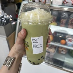 Frothy Matcha Latte - A person holds a large, frothy matcha latte with tapioca pearls in a clear plastic cup with a dome lid. The drink has a light green color and a generous layer of foam on top. The cup has a label indicating it is a frothy matcha latte, size large, with no ice, priced at $8.71. The background shows a store counter with various products and containers.