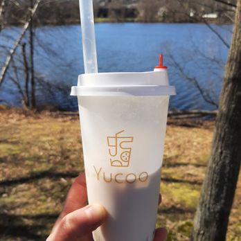 Yucoo - Delicious milk tea with boba - A person holds a white Yucoo-branded milk tea cup with a straw and a sauce bottle, outdoors near a lake. The background features a body of water, trees, and a clear sky, indicating a natural setting. The cup appears to contain a beverage with boba, and the scene suggests a casual outdoor outing. - Bubble Tea Near Me - Brighton, Massachusetts